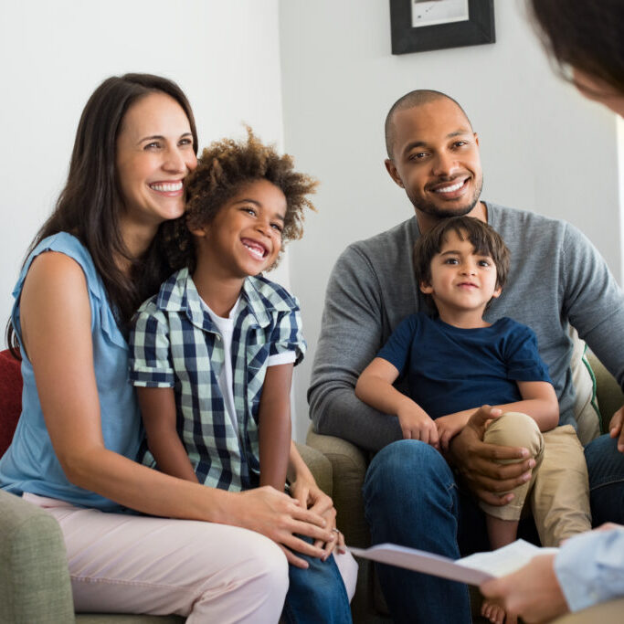 Happy young family sitting on couch and talking with family counselor. Smiling parents with adopted children discussing with counselor. Multiethnic family meeting a financial agent.